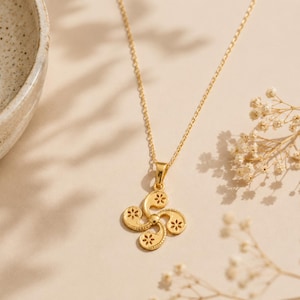 May include: A gold-colored necklace featuring a pendant with a four-armed curved design, known as a Lauburu, with each arm containing a small floral cutout. The pendant hangs from a delicate gold-colored chain link necklace.