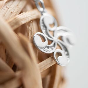 Op de afbeelding: Een zilverkleurige hanger met een spiraalvormig ontwerp met vier armen, voorzien van gladde, gepolijste oppervlakken en getextureerde, verzonken delen. De hanger is bevestigd aan een kleine metalen lus aan de bovenkant en toont een symmetrisch, gebogen patroon.