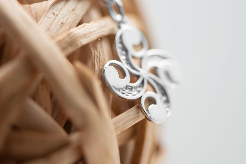 Op de afbeelding: Een zilverkleurige hanger met een spiraalvormig ontwerp met vier armen, voorzien van gladde, gepolijste oppervlakken en getextureerde, verzonken delen. De hanger is bevestigd aan een kleine metalen lus aan de bovenkant en toont een symmetrisch, gebogen patroon.