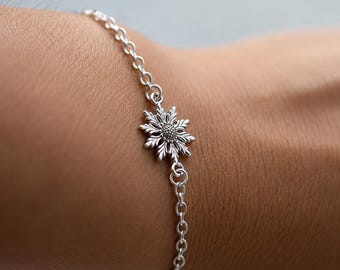 May include: A silver-toned chain bracelet featuring a central metal charm shaped like a stylized flower or snowflake with textured petals and a dotted circular center. The delicate link chain connects to both sides of the floral-shaped metal charm.