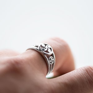 Peut inclure: Une bague en argent avec un design délicat et complexe présentant un motif répétitif de volutes stylisées. La bague est portée à un doigt.