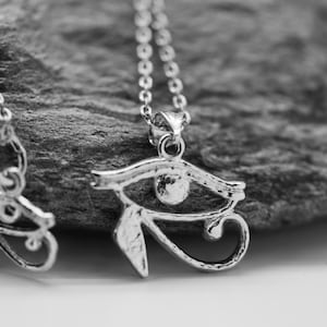 Puede incluir: Dos collares de plata con un colgante de ojo de Horus detallado en una cadena. Los collares están sobre una superficie gris texturizada.