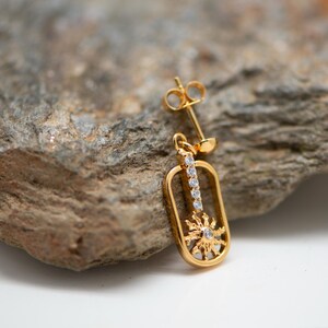 May include: A gold-colored earring with a sunburst design and a row of clear gemstones. The earring is a stud style, with a post for piercing. The earring is set against a textured, gray rock.