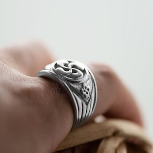 Peut inclure: Une bague en argent avec un motif complexe représentant une fleur stylisée. La bague est portée à un doigt.