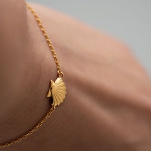 Op de afbeelding: Een gouden kettingarmband met een delicate schelpbedel. De armband zit om een pols.