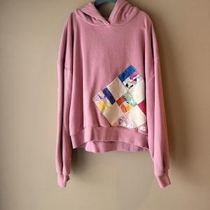 May include: A dusty rose long-sleeve hooded sweatshirt featuring a large, diamond-shaped patchwork panel on the front. The patchwork consists of various colorful fabric squares in shades of blue, yellow, white, and green, stitched onto the garment.