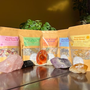 May include: Five brown paper bags with different bath soaks. Each bag has a label with a name and description. The bags are arranged in a row on a wooden surface. There are crystals and a red agate stone in front of the bags.