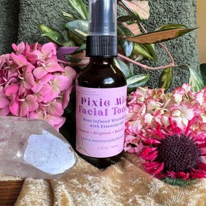 May include: A small glass bottle with a pink label that reads "Pixie Mist Facial Toner". The label also includes the text "Rose infused Witch Hazel with Essential Oils" and "Cleanse - Brighten - Balance". The bottle is sitting on a pink and white flower.