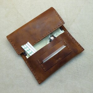 May include: A brown leather cigarette case with a flap closure. The case has a pocket for rolling papers and a slot for a filter. The case is open and there are rolling papers and a filter inside.