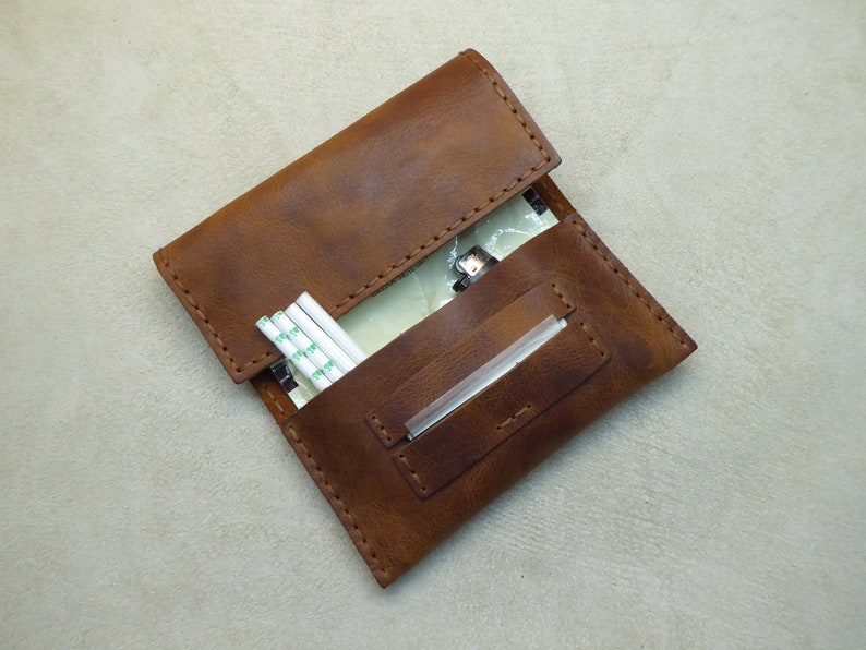 May include: A brown leather cigarette case with a flap closure. The case has a pocket for rolling papers and a slot for a filter. The case is open and there are rolling papers and a filter inside.