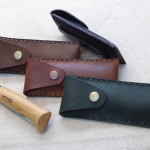 May include: Four leather knife sheaths in brown, dark brown, green, and black. The sheaths have a flap with a brass button closure. A wooden handled pocket knife with a silver blade and the text "Opinel Savoie France" is in the foreground.