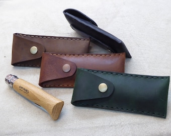 May include: Four leather knife sheaths in brown, dark brown, green, and black. The sheaths have a flap with a brass button closure. A wooden handled pocket knife with a silver blade and the text "Opinel Savoie France" is in the foreground.
