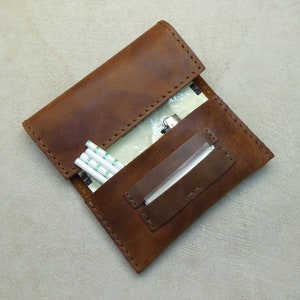 May include: A brown leather cigarette case with a flap that opens to reveal a compartment for rolling papers and a separate compartment for cigarette filters.