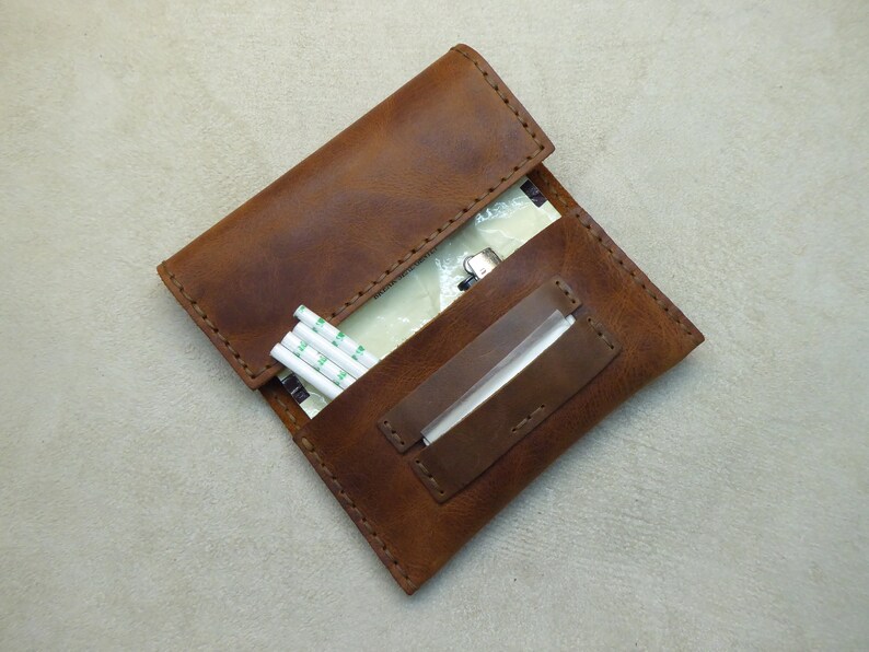 May include: A brown leather cigarette case with a flap that opens to reveal a compartment for rolling papers and a separate compartment for cigarette filters.