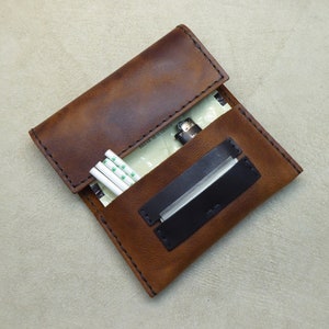 May include: A brown leather cigarette case with a black leather flap and a lighter inside. The case has a stitched edge and a white paper pack of cigarettes inside.