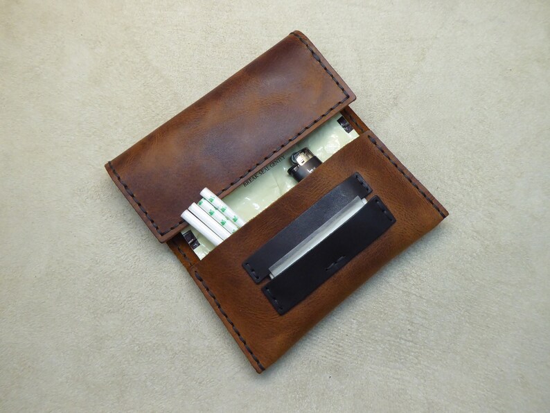 May include: A brown leather cigarette case with a black leather flap and a lighter inside. The case has a stitched edge and a white paper pack of cigarettes inside.