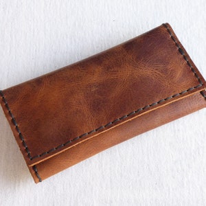 May include: A brown leather key holder with black stitching. The holder is folded closed and has a smooth, textured surface.