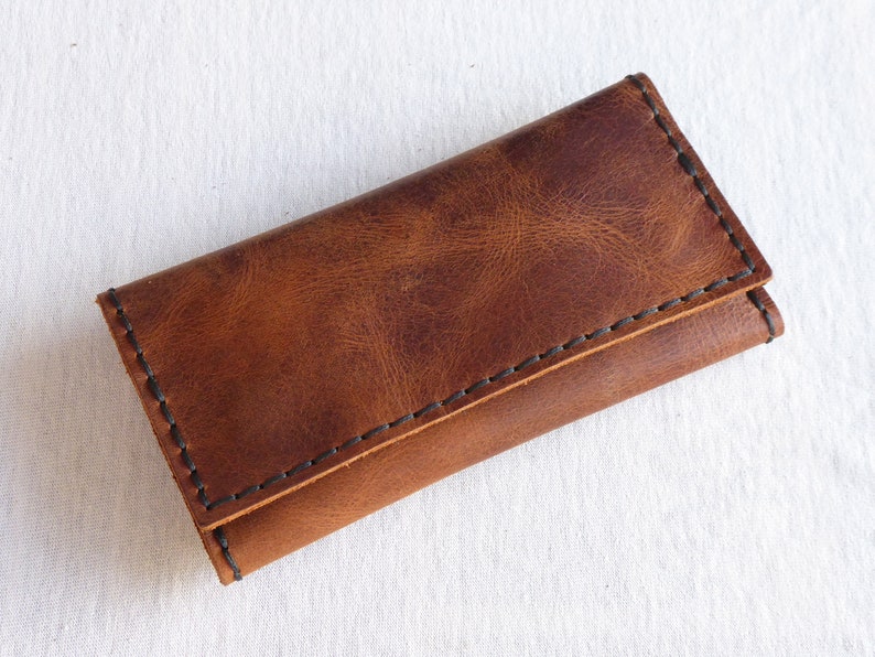 May include: A brown leather key holder with black stitching. The holder is folded closed and has a smooth, textured surface.