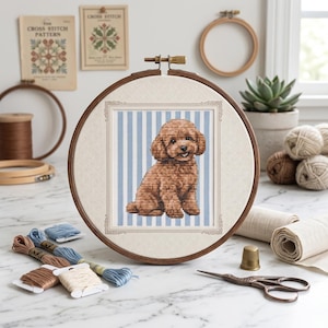 May include: A cross-stitch embroidery project in a wooden hoop featuring a brown poodle puppy sitting against a light blue and white vertical striped background, framed by a decorative border on a cream-coloured fabric canvas.