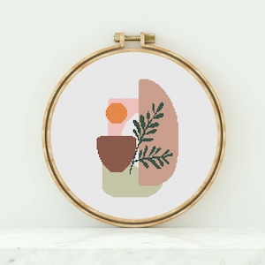 Puede incluir: Un aro de bordado de punto de cruz con un diseño abstracto moderno que presenta formas geométricas en rosa, naranja, marrón y verde.