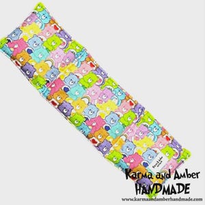 May include: A rectangular, handmade item with a colourful Care Bears pattern. The fabric features various Care Bears in shades of blue, pink, yellow, green, and purple, with rainbows. The item has a white label with the text "Karma and Amber HANDMADE".