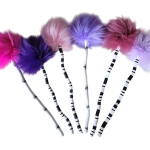 May include: A collection of colorful, fluffy pom-poms on white stems with black and white striped accents. The pom-poms are in shades of pink, purple, and lavender. The stems are curved, creating a whimsical display.
