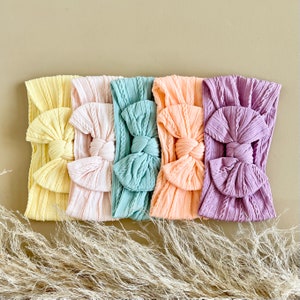 May include: Five fabric headbands in different colours: yellow, pink, light blue, peach, and purple. Each headband has a knotted bow in the centre.