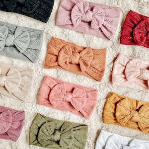 May include: Assortment of knitted headbands with bows in various colours, including beige, brown, pink, green, grey, and red. The headbands are arranged on a white fluffy surface.