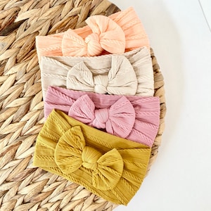 May include: Four fabric headbands with knotted bows in different colours: peach, beige, pink, and mustard yellow. The headbands are made of a textured fabric with a cable knit pattern.