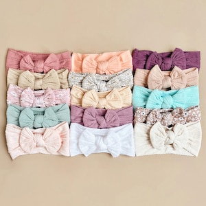 May include: A set of 14 knotted headbands in a variety of colours and patterns, including plain colours, floral prints, and textured fabrics. The headbands are arranged in rows, with each headband featuring a large, knotted bow.