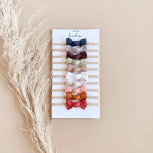 May include: A set of 10 hair bows in various colours, including navy, green, beige, white, pink, orange, and red. The bows are made of fabric and are attached to elastic headbands. The headbands are displayed on a white card with the text "LITTLE LUBA" printed on it.