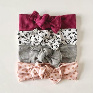 May include: Set of four headbands with different patterns. The headbands are made of a soft, stretchy fabric and feature a knotted bow. The patterns include a burgundy ribbed fabric, a white fabric with black floral print, a grey ribbed fabric, and a pink fabric with brown polka dots.