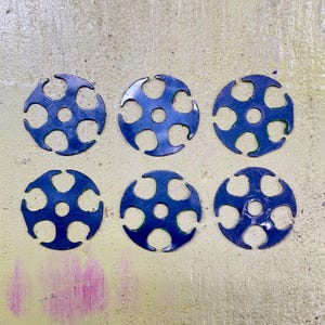 Puede incluir: Seis objetos metálicos azules, circulares, con un diseño similar a una flor. Cada uno tiene un agujero central y seis cortes curvos alrededor del perímetro, creando una apariencia de engranaje. Están sobre una superficie amarilla clara.