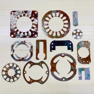 May include: A collection of various rusted metal shapes with circular and geometric designs. The pieces have a distressed appearance with shades of brown, rust, and hints of blue-green. Some shapes have cut-out patterns, and the arrangement is on a light-colored surface.
