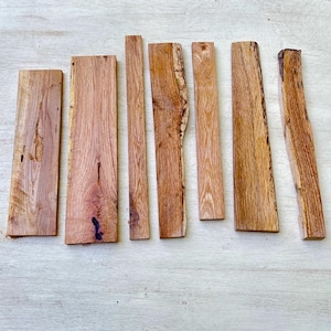 Puede incluir: Un conjunto de siete tablones de madera rectangulares de diferentes anchos y longitudes. Los tablones muestran patrones naturales de vetas de madera y tonos marrones cálidos, algunos con el borde de corteza original. La madera parece sin terminar.