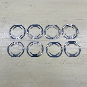 May include: Eight silver-colored metal rings with a unique, open design. Each ring has a circular shape with curved cutouts and small, evenly spaced holes. The rings are arranged in two rows of four on a light-colored surface.