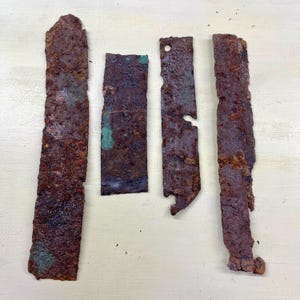 Peut inclure: Quatre pièces de métal rectangulaires rouillées. Les surfaces brunes présentent des zones d'oxydation verte. Les bords sont irréguliers, et une pièce a un trou et une encoche. Longueurs variables.