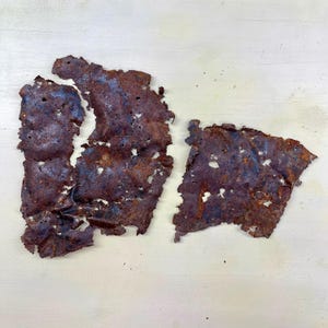 May include: Two irregularly shaped pieces of rusted metal with a dark brown and blue-grey color. The metal has many holes and a rough, textured surface. The pieces are placed on a light-colored wooden surface.