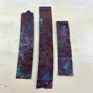 Peut inclure: Trois morceaux rectangulaires de métal vieilli, de couleur brun rouille avec des taches de patine bleu-vert. Les bandes métalliques ont des bords rugueux et inégaux, suggérant qu'elles sont récupérées ou réutilisées.