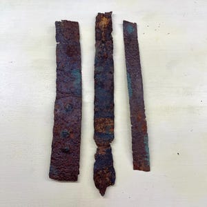 Peut inclure: Trois bandes métalliques rectangulaires rouillées, à surface texturée. Les bandes varient en largeur et longueur, affichant des nuances de brun, de rouille et des reflets bleu-vert. Le métal semble vieilli, suggérant un objet ancien ou récupéré.