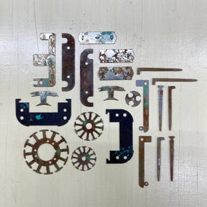 May include: A collection of aged metal hardware components, such as brackets, plates, and nails, displayed on a light surface. The items exhibit signs of wear, with rust and faded paint in hues of blue, green, and brown. The assortment includes diverse shapes and sizes.