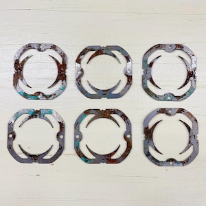 May include: Six weathered, square metal electrical box covers. Each has a gray finish with rust and teal accents. The covers feature a decorative, curved design and small mounting holes, arranged on a white wooden surface.