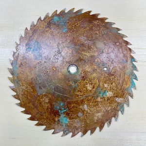 May include: A circular saw blade with a rusty, weathered appearance. The blade has sharp, pointed teeth around the perimeter and a central hole. Patches of turquoise-colored oxidation are visible on the surface, adding contrast to the brown and bronze tones.