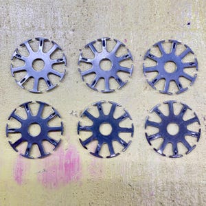 Puede incluir: Seis objetos metálicos circulares plateados, cada uno con un agujero central y radios que terminan en forma de T. Están dispuestos en dos filas de tres sobre una superficie clara. Cada objeto mide aproximadamente 5 cm de diámetro.