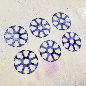 Puede incluir: Seis adornos circulares de metal azul con un diseño similar a una rueda. Cada pieza tiene un agujero central y radios radiales, creando un patrón abierto e intrincado. El metal tiene una apariencia texturizada y ligeramente desgastada.