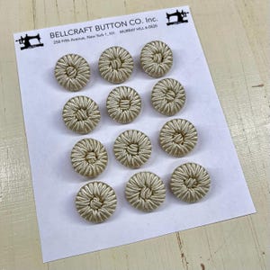 May include: A collection of twelve beige fabric-covered buttons arranged on a white card. The buttons have a circular design with a woven texture. The card includes the text "BELLCRAFT BUTTON CO. Inc."