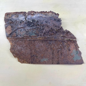 Peut inclure: Une pièce de métal rectangulaire rouillée et corrodée. La surface est principalement brun foncé et noire, avec des zones vertes et bleues. Les bords sont irréguliers et présentent de petits trous.