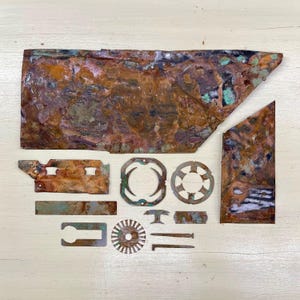 May include: A collection of irregularly shaped, weathered copper pieces with a patina of brown, green, and blue hues. The assortment includes a large rectangular sheet, geometric shapes, and small metal components, likely for crafting or industrial use.