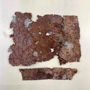 May include: Two irregularly shaped pieces of rusted metal with a mottled, textured surface. The larger piece has a jagged edge and several holes. The color is primarily shades of brown and rust, with hints of blue-gray.