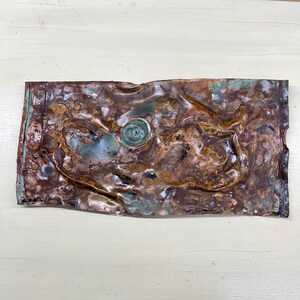 May include: A rectangular, textured metal art piece with a distressed, aged appearance. The artwork features a mix of brown, copper, and teal colors, with a central circular design. The edges show rivet holes.